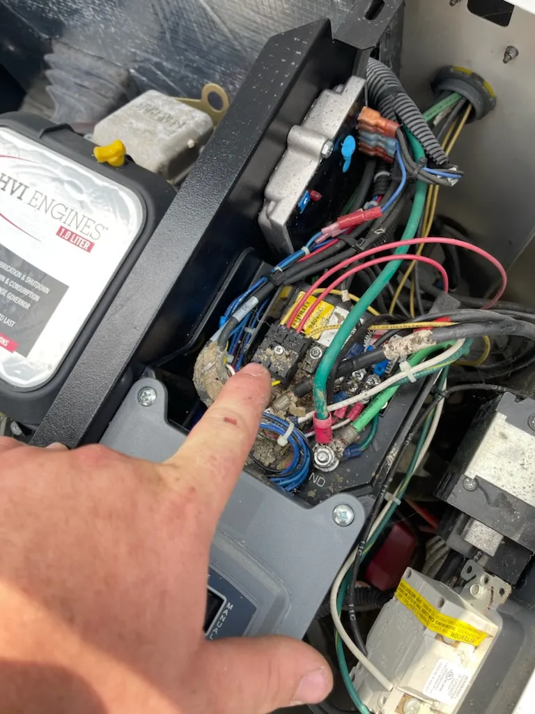 A person points to a component inside an electrical panel containing various wires and connectors near an engine compartment, highlighting the need for professional electrical repair.