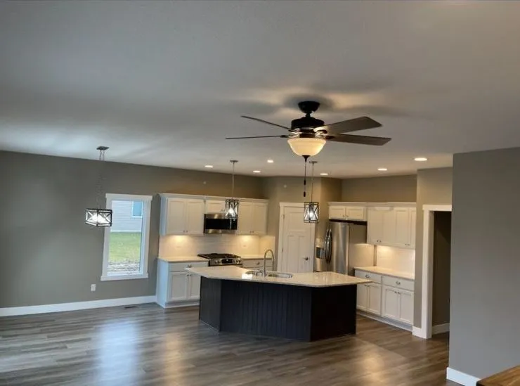 Modern kitchen with white cabinets, stainless steel appliances, center island, pendant lights installed by a skilled electrical contractor, and an adjacent open living space with wood flooring and ceiling fan.
