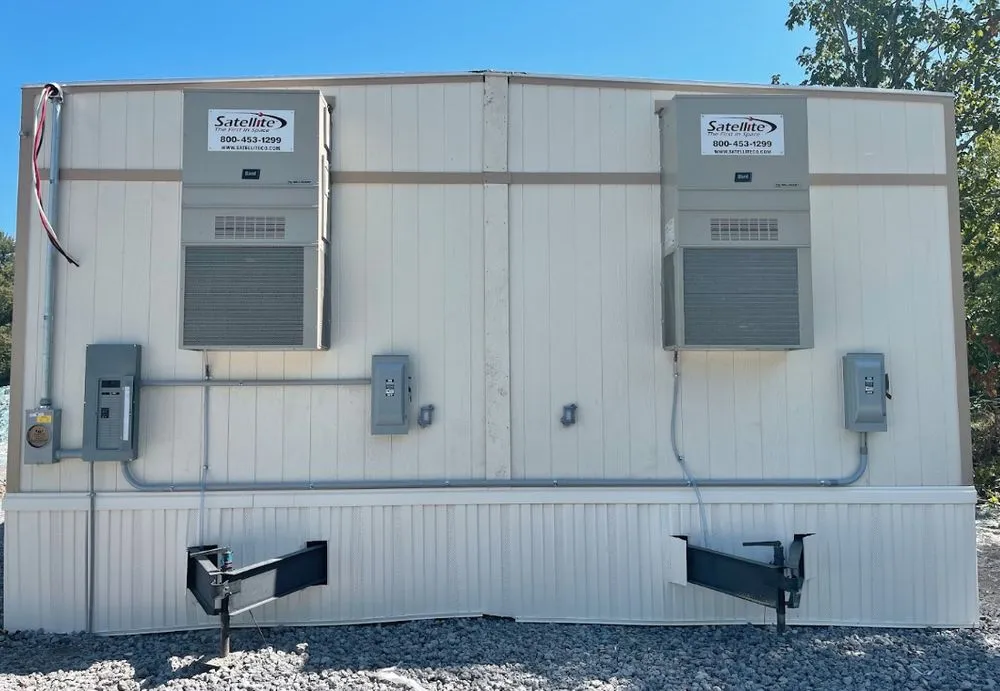 A white portable building with two Satellite air conditioning units mounted on the wall and two trailer hitches at the base sits on gravel, ideal for electrician service in Angola or providing on-site electrical repair.