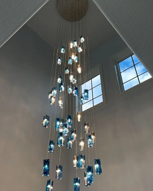A modern chandelier with multiple hanging blue and clear glass pendant lights is suspended in a tall room with two high windows letting in daylight.