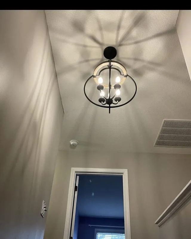 A ceiling light fixture with four bulbs casts intricate shadows on a white ceiling above a hallway with an open door and vent visible.