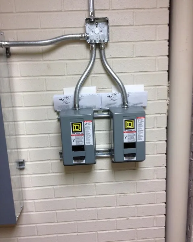 Two gray electrical switch boxes are mounted on a beige brick wall with two pieces of paper partially trapped behind them.