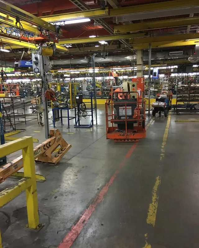 Factory floor with various industrial equipment, machinery, safety railings, and a small orange scissor lift. The area is well-lit and organized.