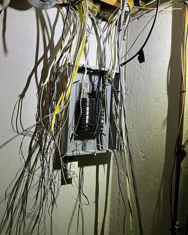 An open electrical panel with numerous tangled wires hanging loosely from the ceiling and wall in a dimly lit room.