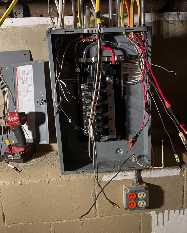 An electrical panel with exposed wires and breakers is mounted on a basement wall, with various cables hanging out and tools placed nearby.