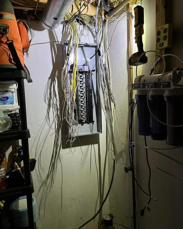 An open electrical panel with numerous loose, disorganized wires hanging down in a dimly lit basement, with various tools and equipment nearby.