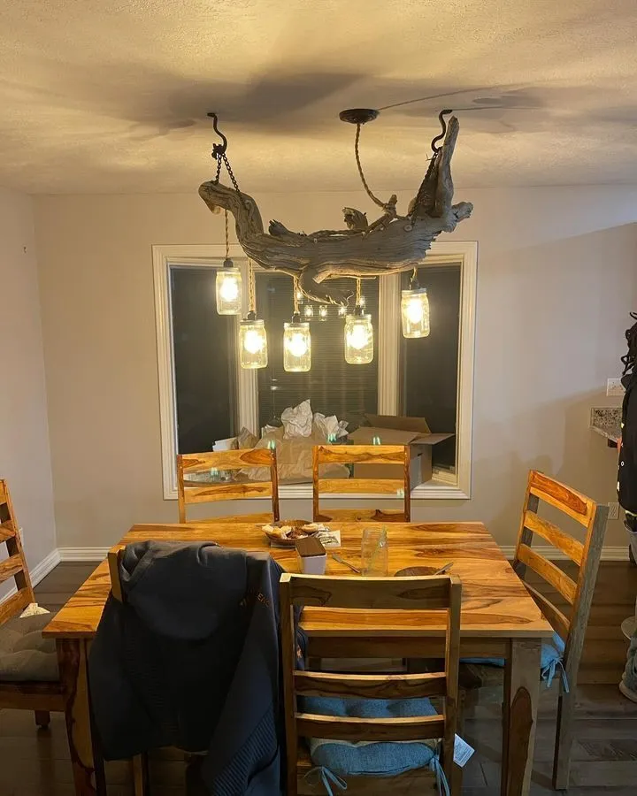 A wooden dining table with six chairs is under a chandelier made from a large piece of driftwood and hanging glass jar lights. The room has a large window and scattered items on the table and floor.