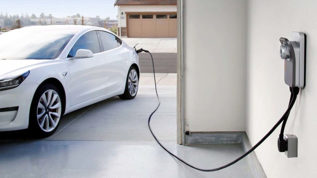 A white electric vehicle is parked in a garage, plugged into a wall-mounted EV charging station with a cable.