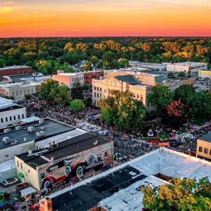 Aerial view of a downtown area at sunset, showing a crowd gathered near a historic courthouse, surrounding buildings, and signage for an Electrician Service in Angola offering expert electrical repair.