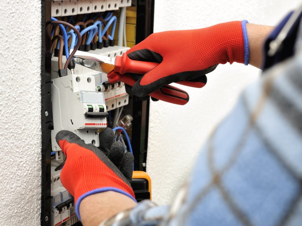 Person wearing red gloves uses pliers and a screwdriver to work on an electrical panel with circuit breakers and wires, showcasing skilled Electrical Repair often needed during Electrician Services in Huntertown.
