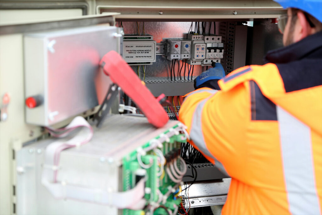 A worker in a high-visibility jacket and blue helmet inspects or repairs electrical components inside an open control panel, showcasing the expertise provided by Electrician Services in Coldwater.