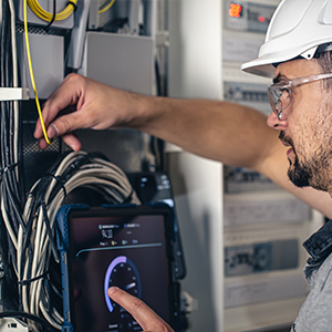 A technician from About All Wire Electric, wearing a hard hat and safety glasses, adjusts cables in a server cabinet while using a tablet displaying a gauge.