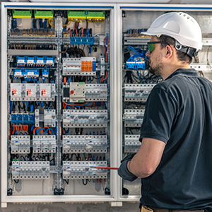 A person wearing a white hard hat and safety glasses inspects or works on an open electrical control panel filled with various switches, wires, and components—demonstrating the expertise of About All Wire Electric.