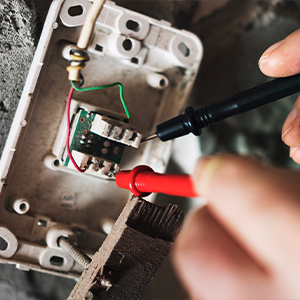 A person from About All Wire Electric tests electrical wires in a wall-mounted socket box using a voltage tester with black and red probes.