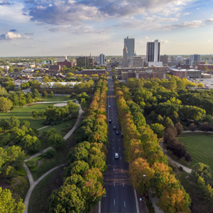 Aerial view of a tree-lined road dividing a city park, with cars on the street and tall buildings in the background, where electrical contractor vans provide reliable electrical services under a partly cloudy sky.