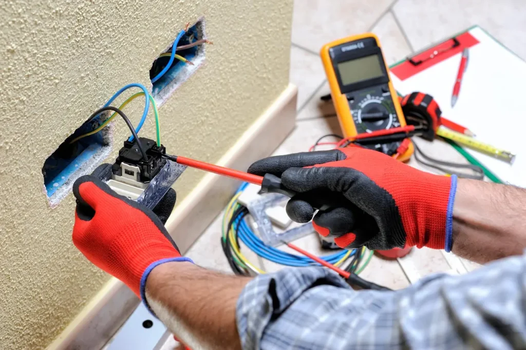 A person wearing red and black gloves installs electrical wiring into a wall socket using a screwdriver, showcasing professional Electrical Repair with tools and a multimeter nearby—ideal for those seeking electrician services in Huntertown.