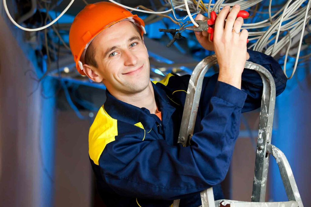 Man wearing an orange hard hat and blue work uniform stands on a ladder, using pliers to work on electrical wiring overhead—perfectly representing professional electrician services in New Haven.