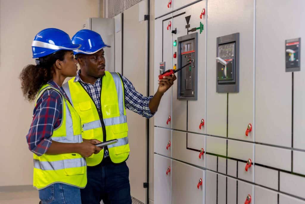 Two engineers wearing safety helmets and reflective vests inspect control panels in an industrial facility, ensuring top-notch electrical repair and maintenance.