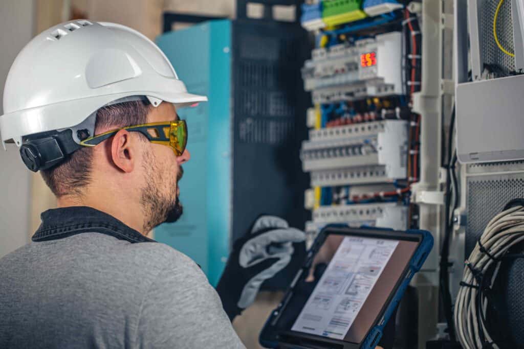 A technician wearing a hard hat and safety glasses uses a tablet while inspecting electrical control panels, exemplifying the professionalism of electrician services in Auburn.