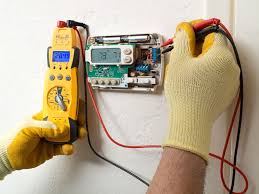 A person wearing yellow gloves uses a digital multimeter to test the wiring of an exposed electronic thermostat on a wall, showcasing professional electrical repair skills.