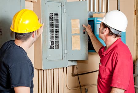 Two workers in hard hats inspect an open electrical panel in a utility room, discussing necessary electrical repair. One points at the panel’s interior while the other observes, highlighting professional Electrician Services in New Haven.