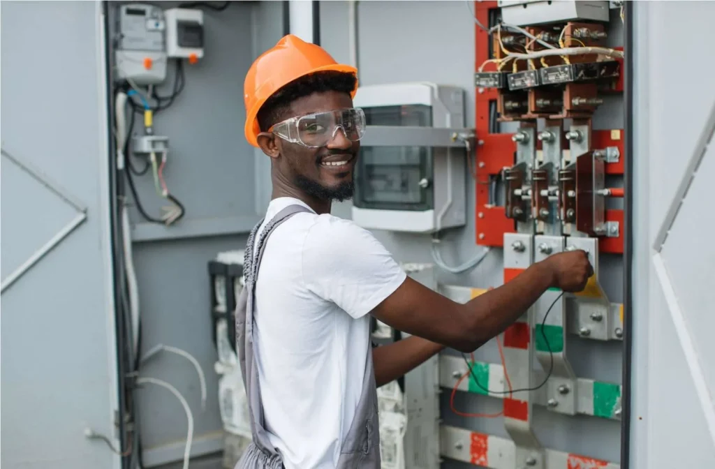 A man wearing a hard hat, safety glasses, and overalls works on an electrical panel, smiling at the camera—showcasing expert electrician services in Fort Wayne.