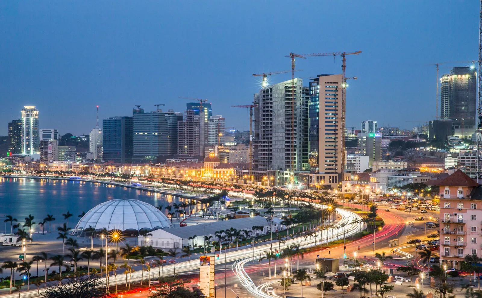 A cityscape at dusk shows tall modern buildings, cranes, a lit coastline, a dome structure, and light trails from traffic on a busy curved road—highlighting the vital role of electrical services in powering urban life.