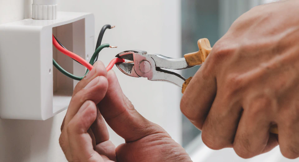 A person uses pliers to cut a red electrical wire protruding from a wall-mounted electrical box, demonstrating electrical repair alongside black and green wires.