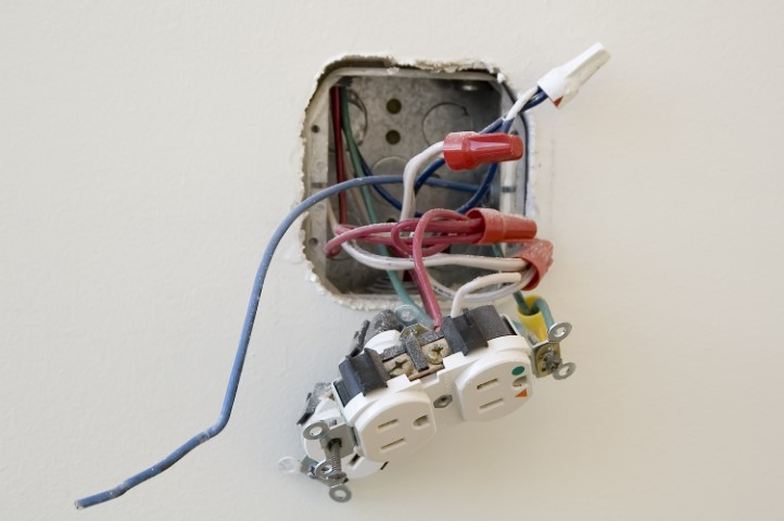An electrical outlet removed from the wall exposes multiple colored wires and wire connectors, revealing electrical work in progress. Ensuring proper electrical safety is essential during installations or upgrades like GFCI or AFCI outlets.